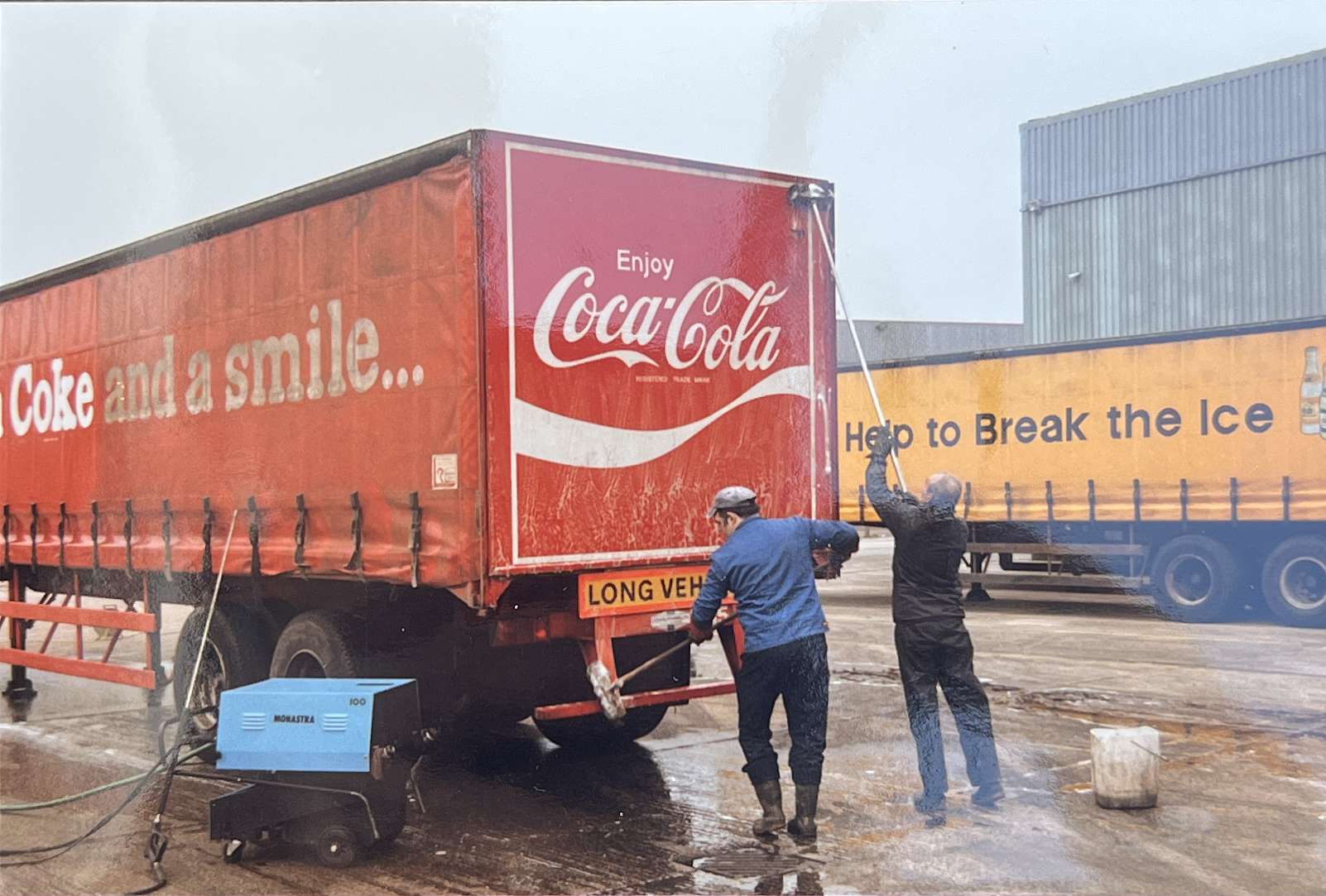 Two workers manually washing a red Coca-Cola haulage trailer.
